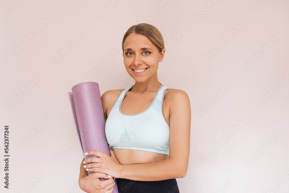 A young caucasian smiling fit tanned blonde woman holding yoga mat in her hands isolated on a light beige color background. Fitness, workout concept. Healthy lifestyle, wellness, sports A young caucasian smiling fit tanned blonde woman holding yoga mat in her hands isolated on a light beige color background. Fitness, workout concept. Healthy lifestyle, wellness, sports