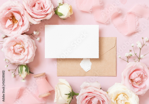 Card with envelope between pink roses and pink silk ribbons on pink top view, wedding mockup