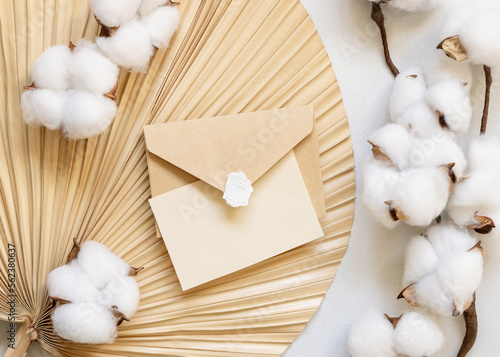 Blank card and envelope on dried palm leaf with cotton flowers top view, wedding mockup