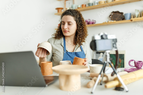 Females potter making a vlog, recording online course, lessons in her studion with earthenware shelf on background