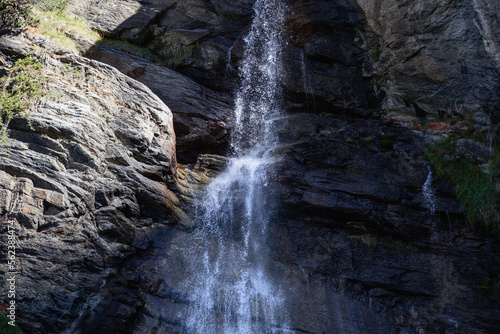 Splashes from falling water of Lillaz waterfall  Cascate di Lillaz  break on granite ledges of alpine rocks  sparse vegetation on sunny side  Aosta valley  Italy