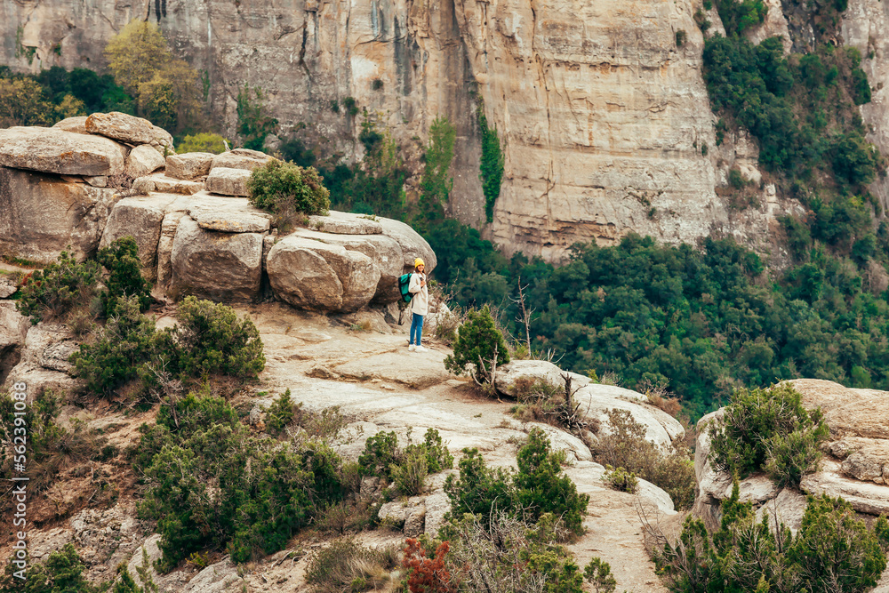 Young girl wears yellow beanie and hiking bag stands on the shore of a mountain lake, looks at camera. Landscape portrait. Outdoor sports, adventure, lifestyle, travel, vacations, holiday concept. Young girl wears yellow beanie and hiking bag stands on the shore of a mountain lake, looks at camera. Landscape portrait. Outdoor sports, adventure, lifestyle, travel, vacations, holiday concept.