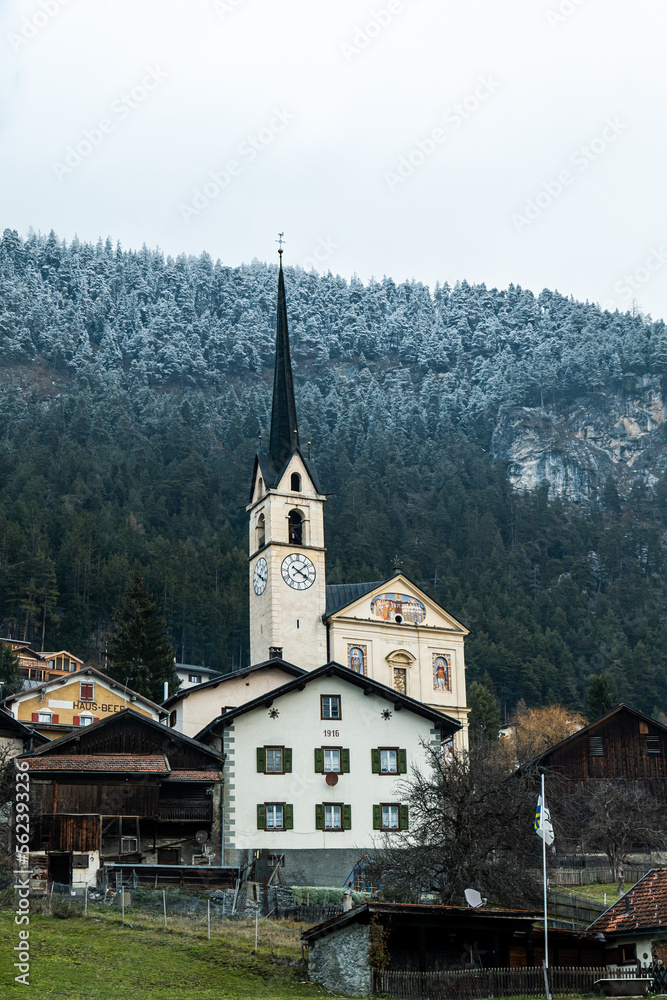 Church surrounded by traditional Swiss houses beneath a mountain with snow covered trees on top in Switzerland.