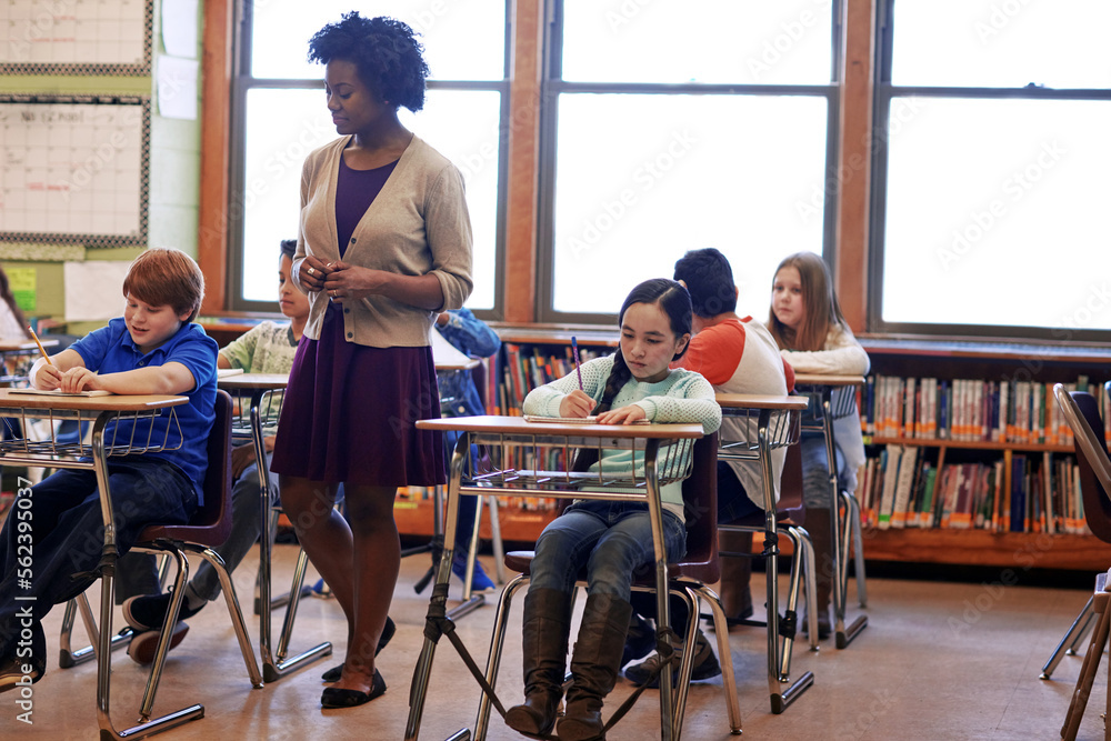 Kids, classroom and teacher woman at school, focus and education for development, learning and future. School kids, black woman and studying at table in class, diversity and notebook for writing test Kids, classroom and teacher woman at school, focus and education for development, learning and future. School kids, black woman and studying at table in class, diversity and notebook for writing test