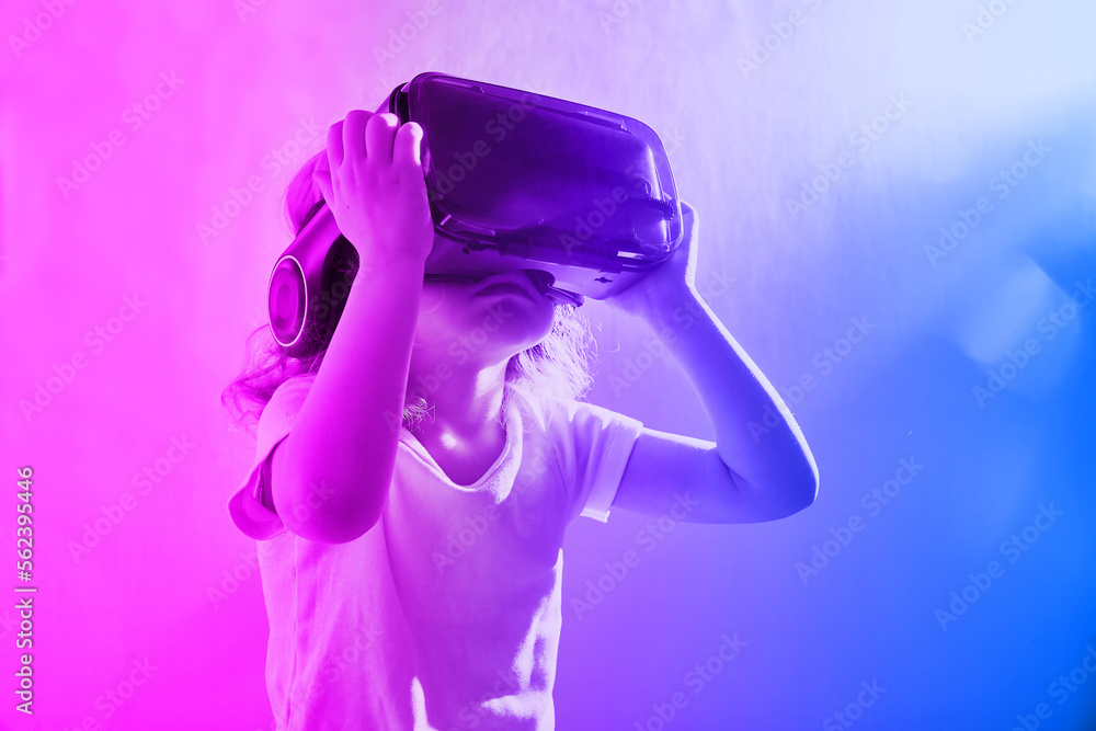 Cute curly-haired girl looks interested in VR glasses. Neon light on the background with copy space Cute curly-haired girl looks interested in VR glasses. Neon light on the background with copy space