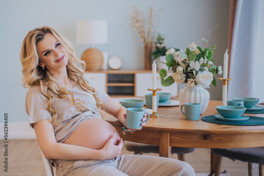 Adorable blonde Scandinavian pregnant woman sitting at kitchen table smiling holds cup of tea at home. Healthy caucasian woman with long wavy hair relaxing, pregnancy. Satisfied female dreaming Adorable blonde Scandinavian pregnant woman sitting at kitchen table smiling holds cup of tea at home. Healthy caucasian woman with long wavy hair relaxing, pregnancy. Satisfied female dreaming