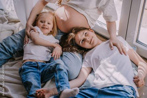 Εκτύπωση καμβά Cropped shoot of two cheerfull sisters in blue jeans and white t-shirts laying on the floor close to pregnant moms belly looking at camera