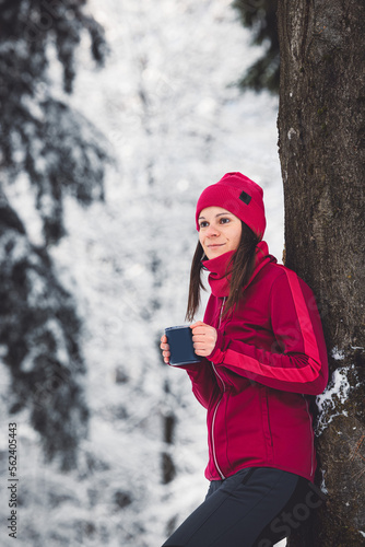 Portrait of beautiful woman in red jacket leaning against a tree in the snowy forest holding a metal reusable cup with hot tea in it