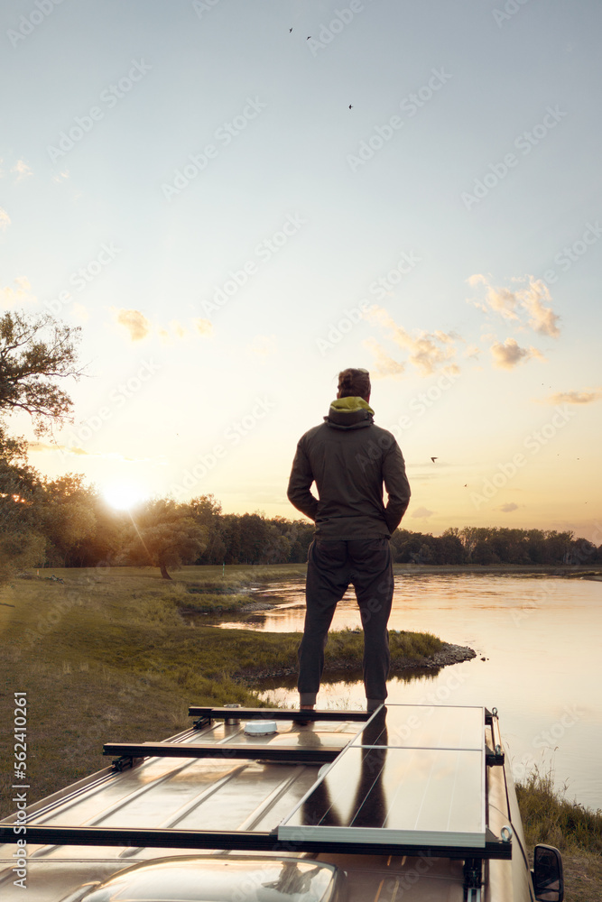 Person standing on top of a camper van next to a river at sunset