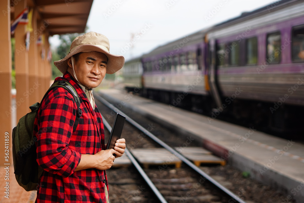Asian man traveller is at railway station, wears hat, holds smart tablet. Concept, travel by train  in Thailand can book or by ticket online. Technology and transportation.                 