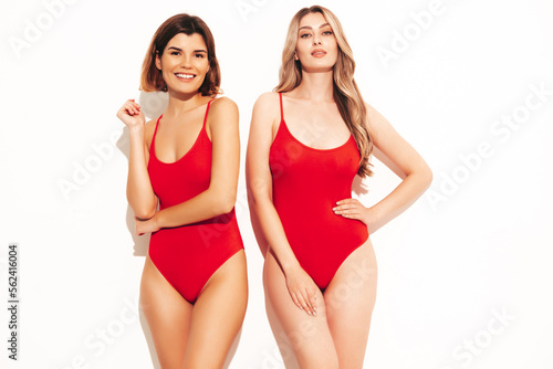 Two beautiful sexy smiling hipster women in red summer swimwear bathing suits. Trendy models having fun in studio. Hot female isolated on white. Cheerful and happy. Slim and fit