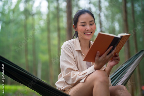 Asian woman relax and enjoy outdoor lifestyle camping in forest mountain on summer holiday travel vacation. Attractive girl resting on hammock and reading a book in pine tree forest in the morning
