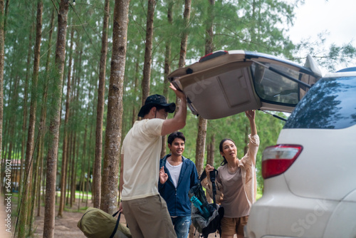 Group of Asian people friends enjoy outdoor lifestyle road trip and camping together on summer holiday travel vacation. Man and woman taking off camping supplies from car trunk at natural park.