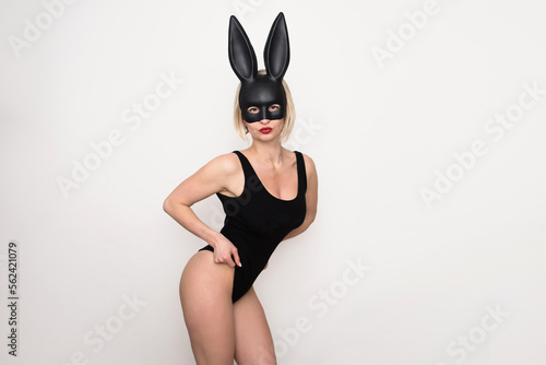A sexy blonde woman dressed in a black Easter bunny costume and bunny ears stands on a white background and poses sensually. Halloween and Easter Bunny Costume - Hot Girl.