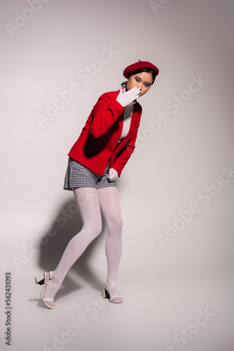 Stylish asian woman in red beret and gloves holding hand near mouth on grey background