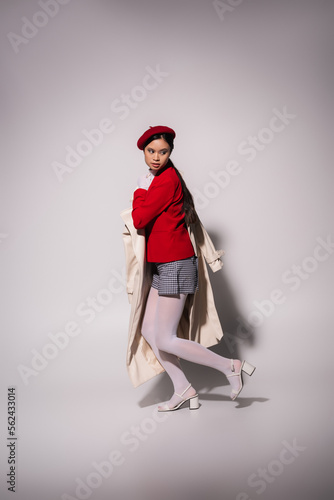 Full length of stylish asian woman in red jacket and beret holding trench coat on grey background © LIGHTFIELD STUDIOS