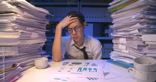 Stressed asian business man is sitting at desk covered with stack of paperwork and having nervous breakdown while working hard at late night. Angry alone male is frustrating and  throws paper in air.