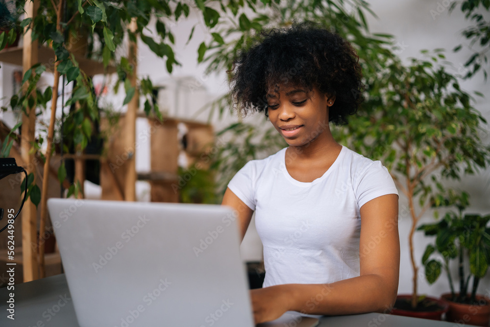 Close-up of smiling African-American businesswoman remote working sitting at desk in home office room with modern biophilia design, on background of green plants. Concept of house garden.