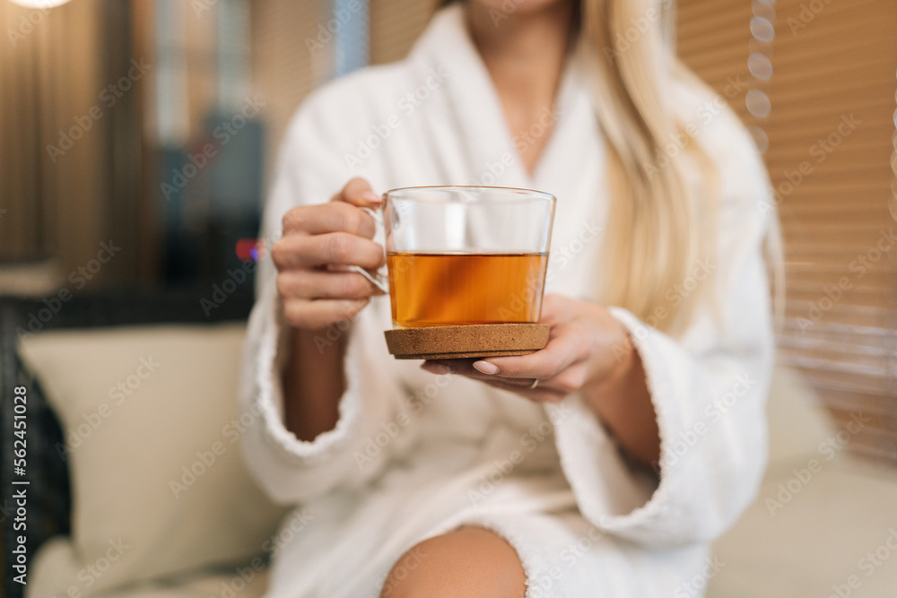 Close-up cropped shot of unrecognizable young woman in white bathrobe holding cup with fresh herbal tea, sitting on sofa in spa salon after treatment procedures. Concept of luxury professional massage Close-up cropped shot of unrecognizable young woman in white bathrobe holding cup with fresh herbal tea, sitting on sofa in spa salon after treatment procedures. Concept of luxury professional massage
