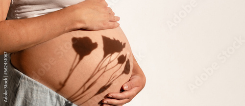 Cropped shot of pregnant woman touching,caressing beautiful big tummy,belly on white background.Female hand on baby bump, flower shadow.Beautiful sunlight, happy pregnancy in spring, summer.Copyspace.