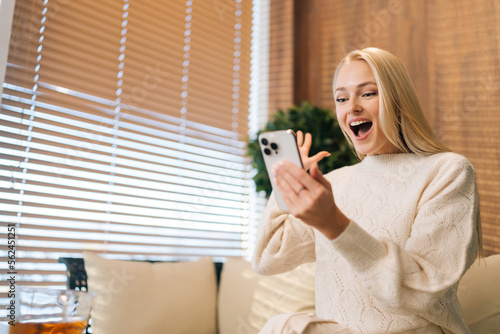 Low-angle view of joyful young woman using smartphone for video call sitting on sofa. Smiling blonde female pleasant online meeting sitting on couch at home, enjoys online chatting with friends