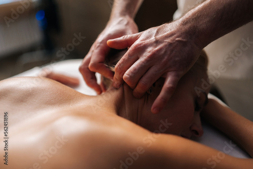 Close-up hands of unrecognizable male massage therapist gently massaging neck of woman with beautiful skin and shiny from massage oil, lying on massage table in spa salon with dark interior.