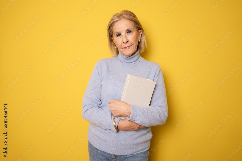 Photo of mature woman happy positive smile holding a book isolated over yellow background Photo of mature woman happy positive smile holding a book isolated over yellow background
