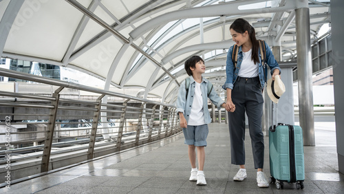 Family Trip Concept. Portrait of a cheerful Asian boy having fun and holding hands with his mother, ready for vacation, standing on the luggage cart. Parents walking with baggage enjoy a vacation.