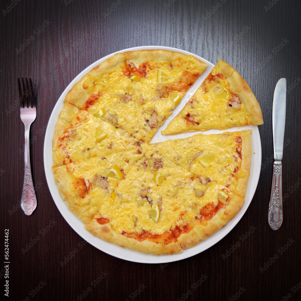 Delicious fresh round cheese pizza with fish, lemon on black table, on white plate, fork, knife nearby, top view Delicious fresh round cheese pizza with fish, lemon on black table, on white plate, fork, knife nearby, top view