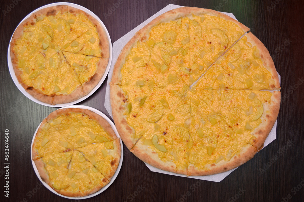 Three delicious fresh round cheese pizzas with pear, pineapples on a black table, surface, on a white plates, three different sizes for serving in a pizzeria Three delicious fresh round cheese pizzas with pear, pineapples on a black table, surface, on a white plates, three different sizes for serving in a pizzeria