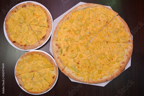 Three delicious fresh round cheese pizzas with pear, pineapples on a black table, surface, on a white plates, three different sizes for serving in a pizzeria