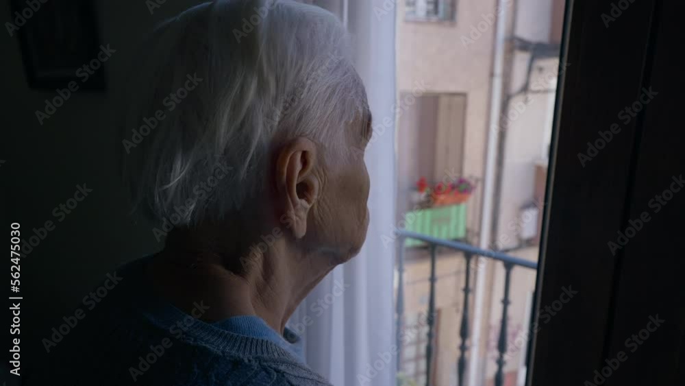 Close-up grey-haired wrinkled old woman standing in darkness looking out window. Unrecognizable Caucasian female senior retiree admiring sunny summer street standing at home indoors Close-up grey-haired wrinkled old woman standing in darkness looking out window. Unrecognizable Caucasian female senior retiree admiring sunny summer street standing at home indoors