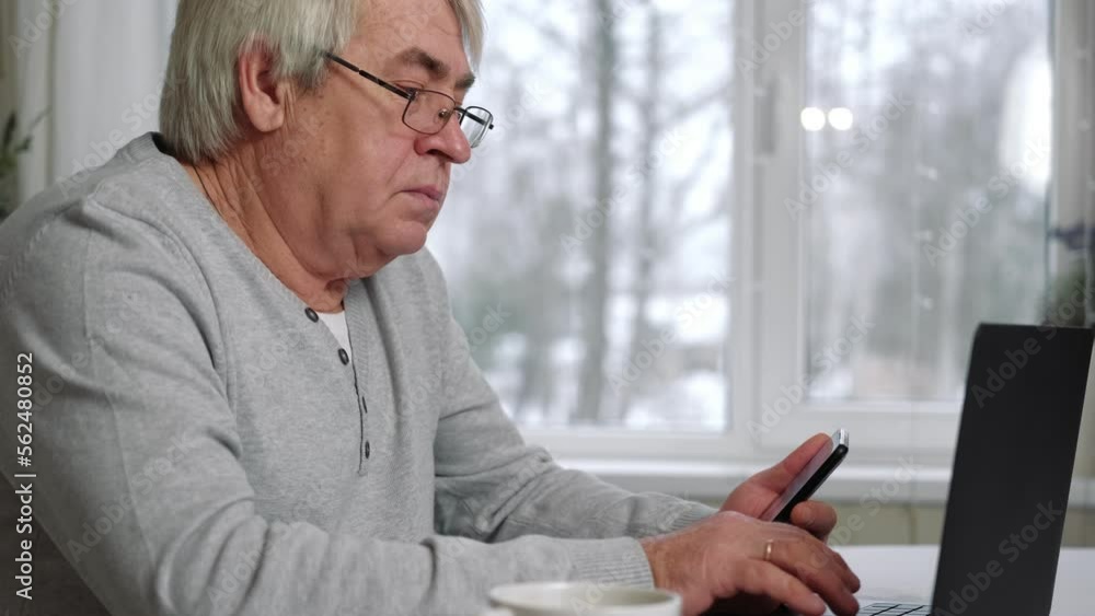 Handsome serious senior man wearing glasses using mobile phone while sitting at desk at his cozy workplace with laptop. Retired male working from home chatting with colleagues. Old Man with Technology Handsome serious senior man wearing glasses using mobile phone while sitting at desk at his cozy workplace with laptop. Retired male working from home chatting with colleagues. Old Man with Technology