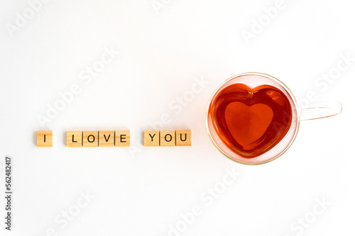 Herzförmige Teetasse aus Glas mit rotem Tee gefüllt auf weißem Hintergrund, Holzplättchen mit den Buchstaben I LOVE YOU links daneben aufgereiht, horizontal, Topview, noch freier Platz  photo