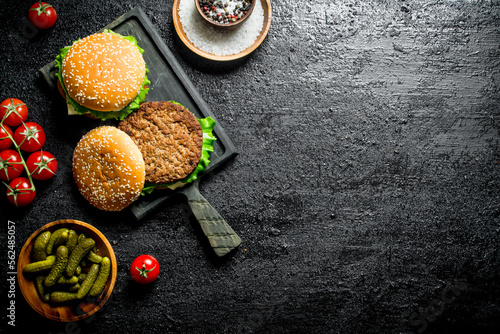 Burgers on a cutting Board with gherkins and spices in bowls.