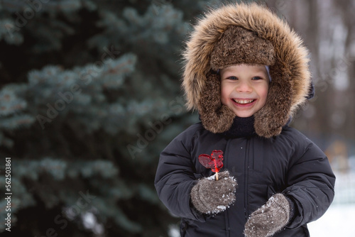 Canvas Print Portrait of a beautiful little Russian boy in a hat with earflaps with a lollipop cockerel in winter in the park