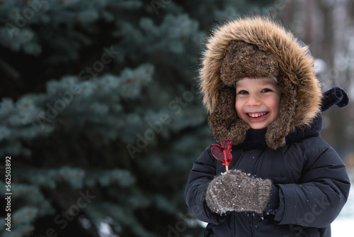 Portrait of a beautiful little Russian boy in a hat with earflaps with a lollipop cockerel in winter in the park Fototapeta