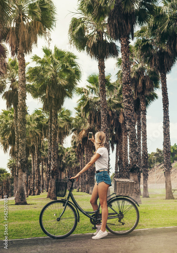 Young blond girl with long hair is on a bycicle and taking a picture of a nice palms alley.