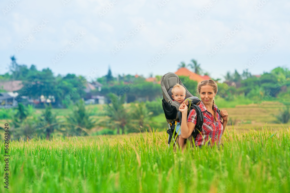 Nature walk in green rice terrace field. Happy mother hold little traveller in carrying backpack. Baby ride on woman back. Travel adventure, hiking with child carrier, family summer vacation on Bali.