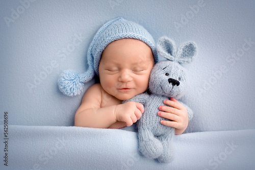 A cute newborn boy in the first days of life sleeps naked on a blue fabric background. The kid gently hugs a light blue knitted bunny. Studio professional macro photography, newborn baby portrait.