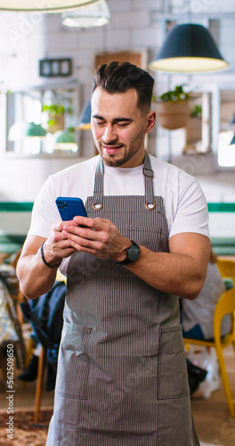 Portrait of happy young handsome Caucasian guy texting on smartphone standing in coffee house in apron at work, looking at camera and smiling. Restaurant service, business concept. Vertical video