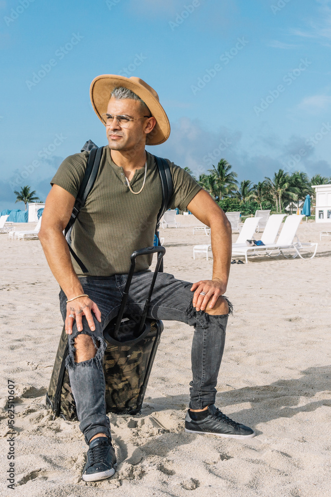 Close up of a traveler handsome hipster man sitting on the carrying baggage, beautiful beach, enjoying amazing view. Wearing a hat and a ripped jeans