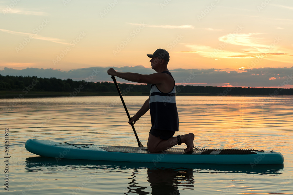 A man on his knees on a SUP board with an oar at sunset swims in the calm water of the lake against the backdrop of a pink sky reflection in the water.