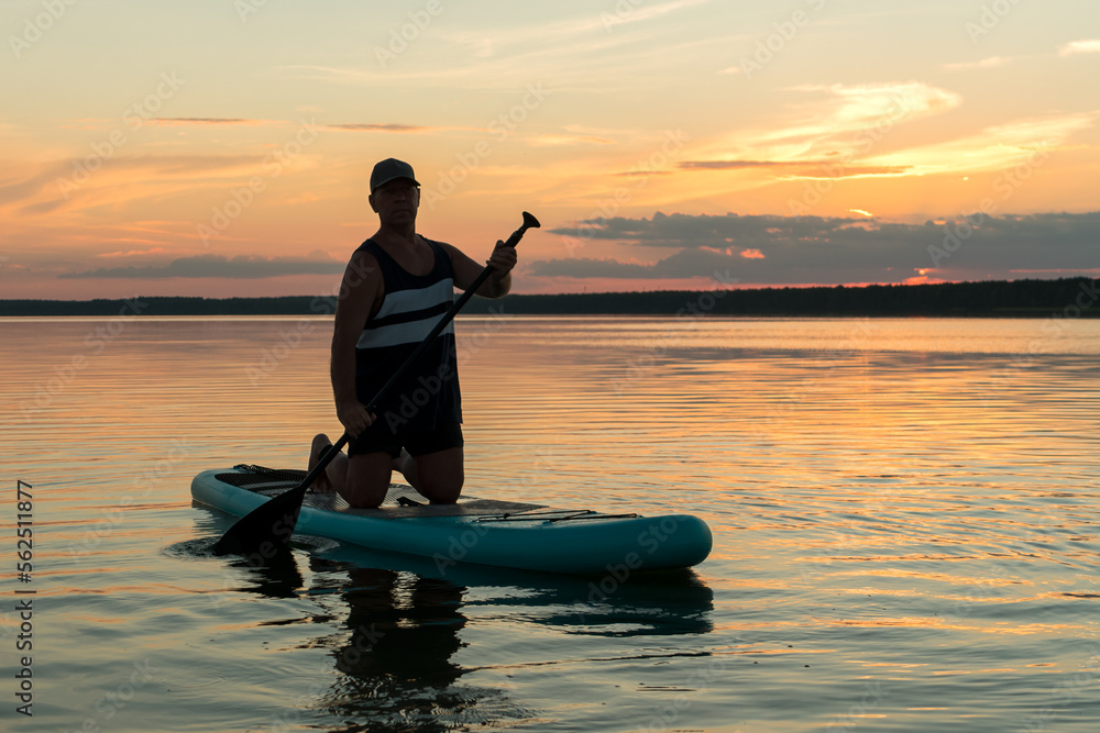 A man in shorts on his knees on a SUP board with a paddle at sunset swims in the water of the lake in the glare from the setting sun.