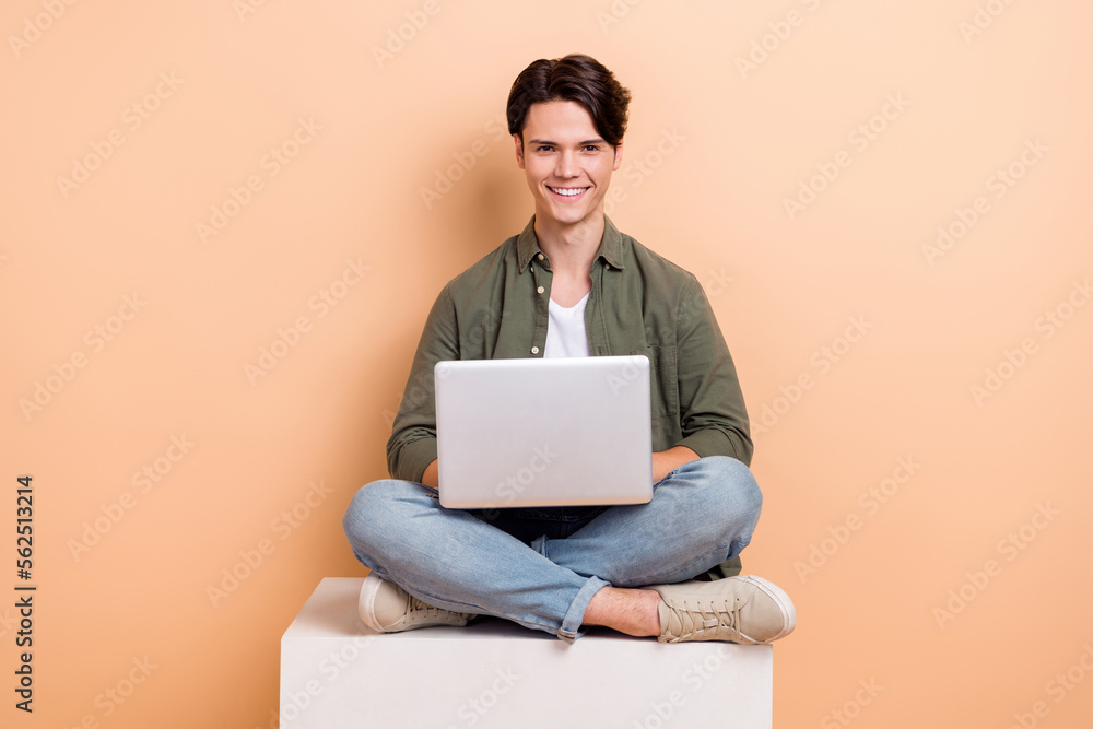 Full length cadre of young optimistic man sitting platform cube use laptop browsing information online education isolated on beige color background Full length cadre of young optimistic man sitting platform cube use laptop browsing information online education isolated on beige color background