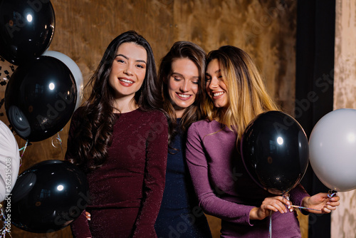 Indoor close up photo of charming happy three girls dressed holiday evening dresses spend time together, dancing and celebrating birthday with balloons 