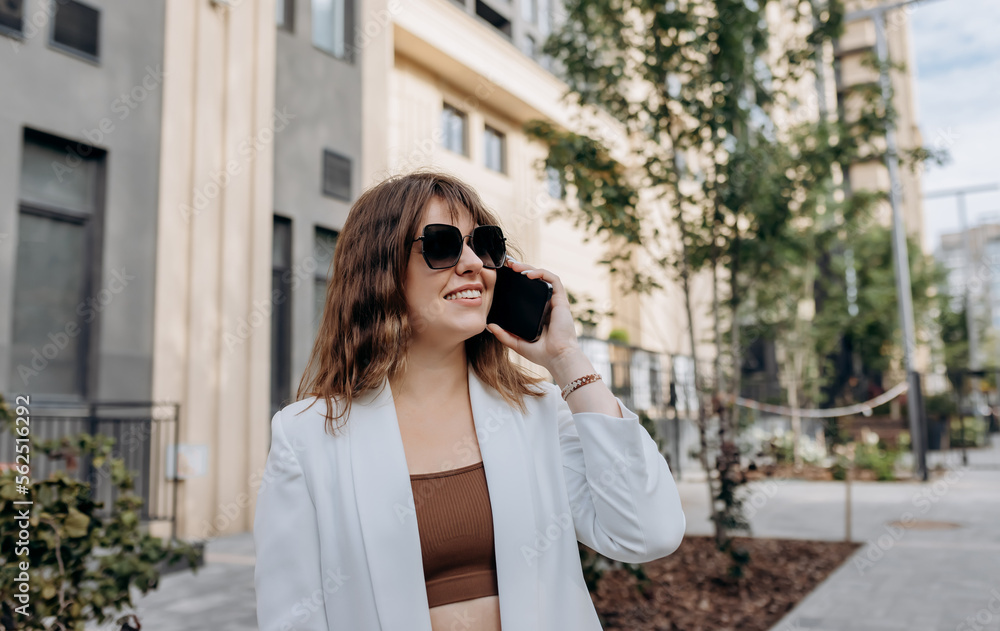Smiling businesswoman in white suit talking phone during walking in city with modern architecture Smiling businesswoman in white suit talking phone during walking in city with modern architecture