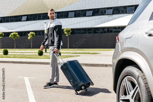 Attractive young man with a suitcase on wheels near the car in sunny weather © AvokadoStudio