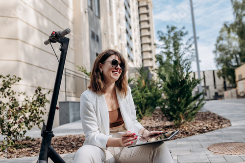 Young businesswoman in white suit sitting on electric scooter and working on digital tablet in city with modern architectury photo