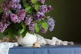 purple lilac in a white jug and a shell on the table on a dark green background.